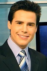 Luiz Bacci em 2007 como âncora do Jornal SBT Rio, de terno escuro, camisa clara e gravata listrada, sorrindo em estúdio com painel ‘Jornal SBT Rio’ ao fundo.