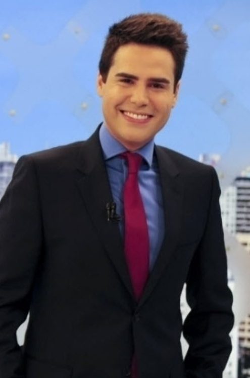 Luiz Bacci em 2010 como apresentador do Balanço Geral SP, com terno preto, camisa azul clara e gravata vermelha, ao lado de tela com logo do programa e cenário de prédios ao fundo.