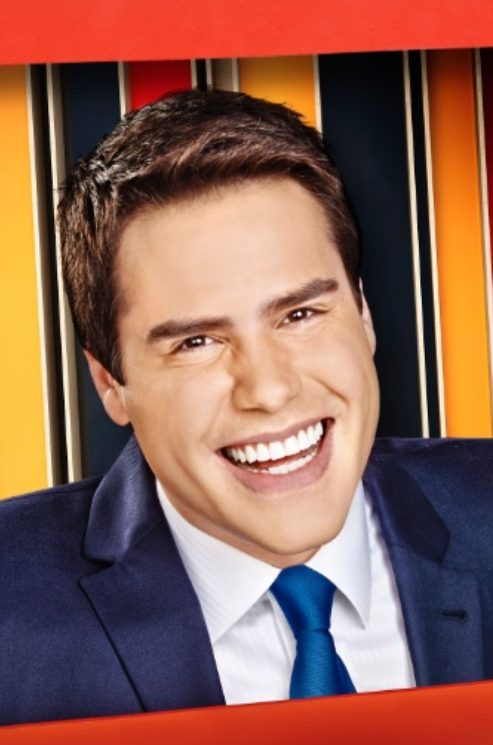 Luiz Bacci em 2014 no programa Tá na Tela da Band: veste terno azul escuro, camisa branca e gravata azul, sorrindo dentro de um frame vermelho, com painéis coloridos ao fundo.