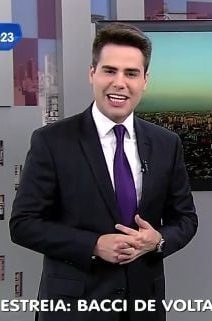 Luiz Bacci em 2015 no telejornal matinal da Record, de terno preto, camisa branca e gravata roxa, apresentando ao vivo com a legenda ‘ESTREIA: BACCI DE VOLTA À RECORD’ e paisagem urbana ao fundo.