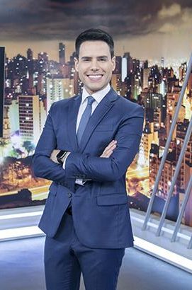 Luiz Bacci em 2017 no estúdio do Cidade Alerta da Record TV, de terno azul escuro, camisa branca e gravata azul, com logo do programa e skyline noturno ao fundo.