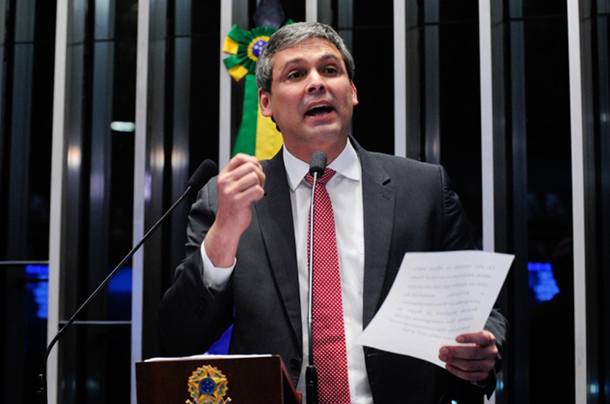 A imagem mostra Lindbergh Farias, líder do PT na Câmara, vestindo terno preto, camisa branca e gravata vermelha com detalhes brancos, falando em um púlpito. Ele segura um papel na mão direita e parece estar fazendo um discurso enfático, com a mão esquerda em gesto de ênfase. 