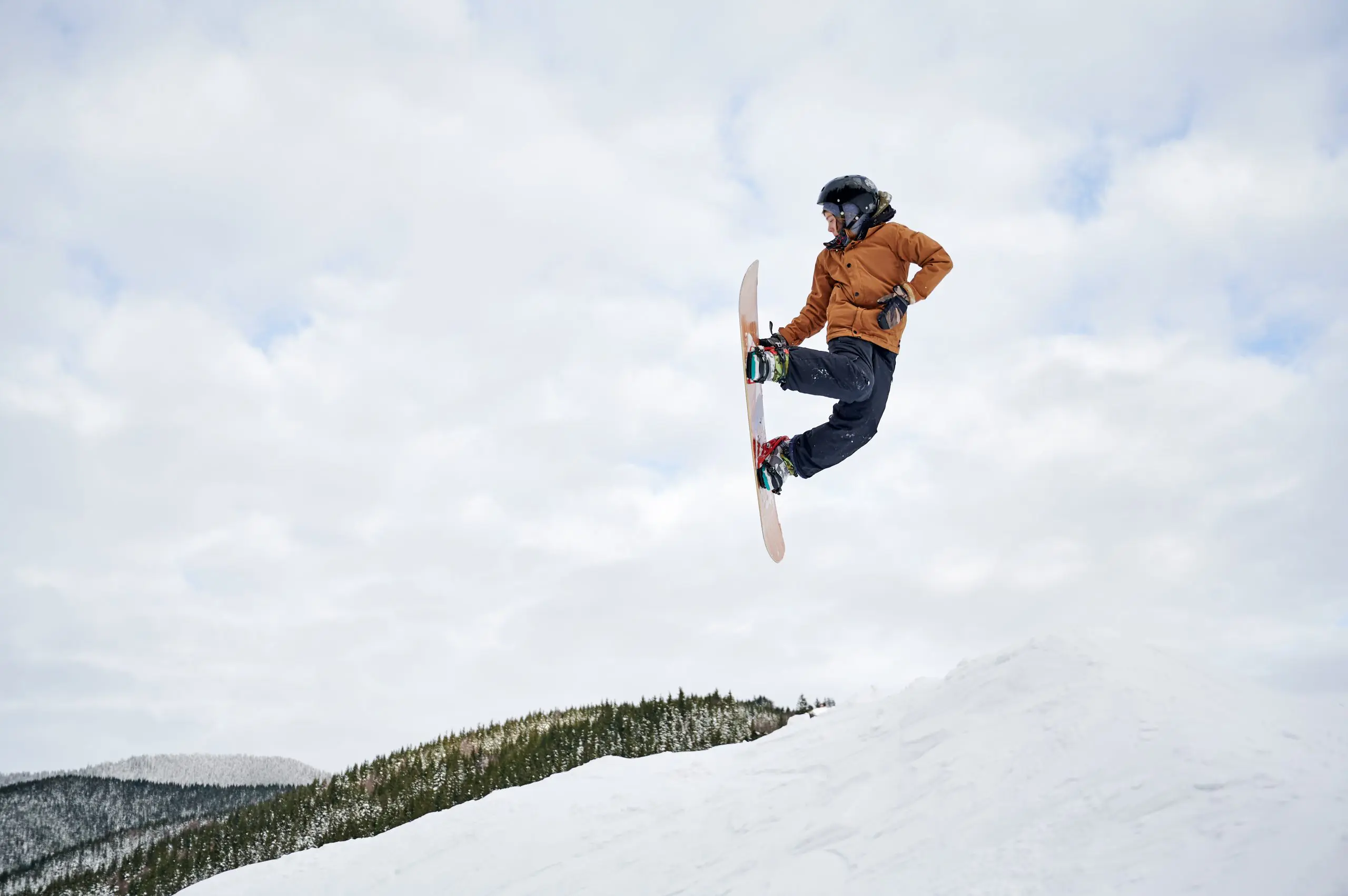 Guy snowboarder in winter jacket and helmet jumping in the air. Adorable kid making jump with snowboard while sliding down snowy hill in winter mountains. Concept of winter sport activities.